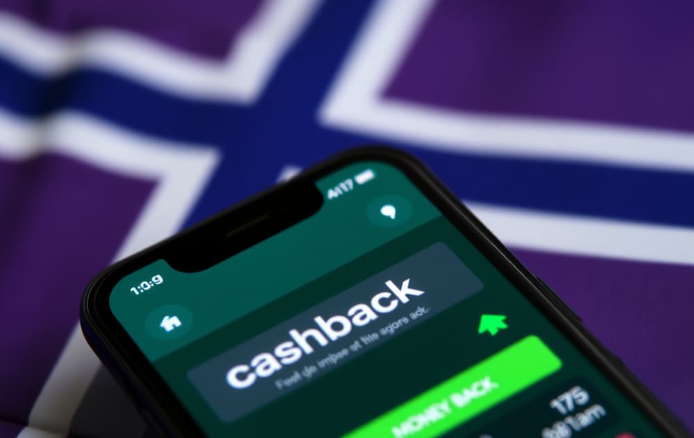 Nærbilde av smarttelefon med betting-app som viser cashback-tilbud, norsk flagg i bakgrunnen og grønn pil som indikerer penger tilbake.
